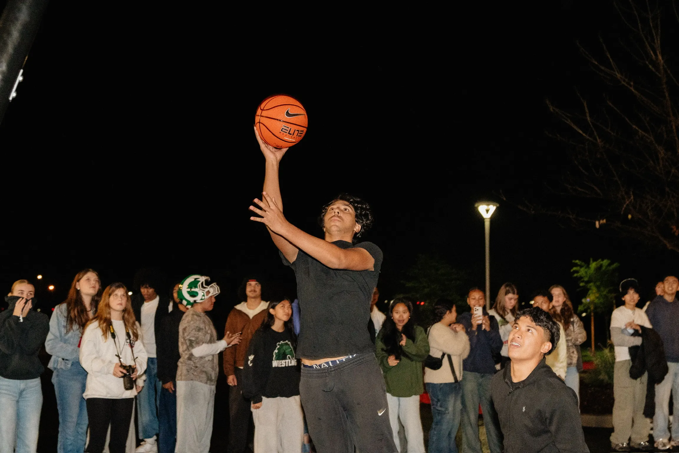 A student leaps for a jump shot during an outdoor night basketball game as a crowd looks on