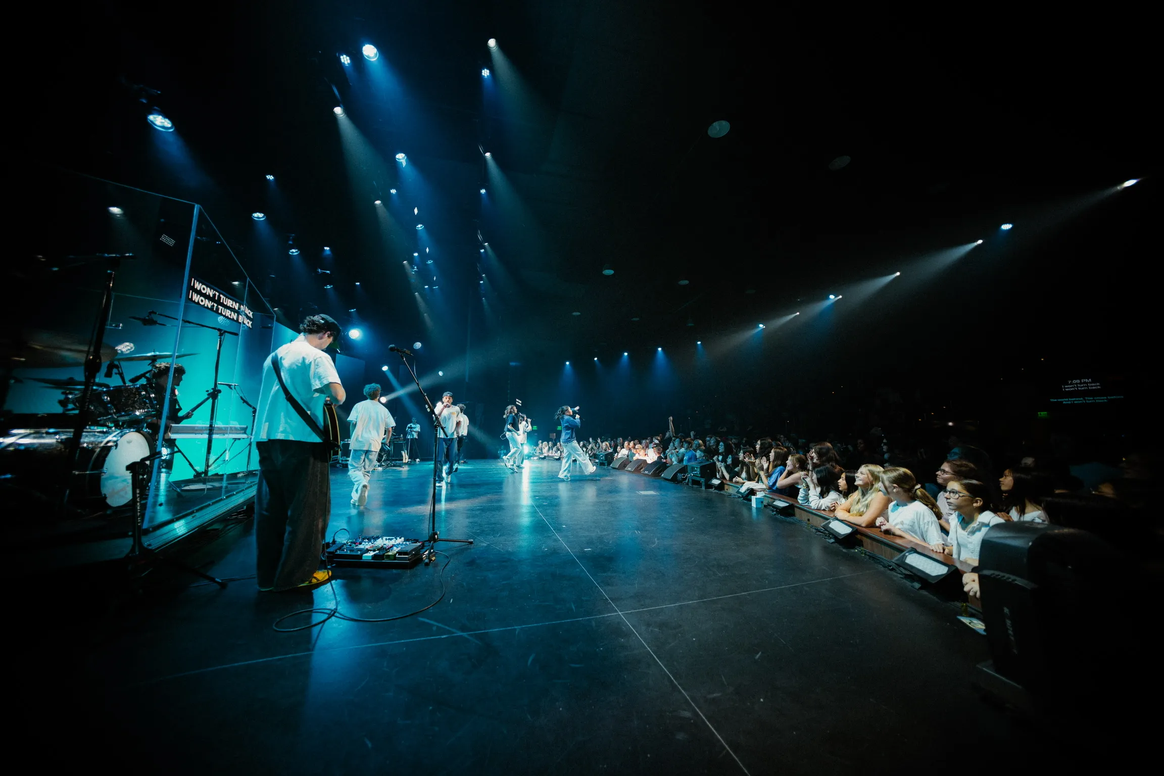 A band performs on stage with a guitarist in the foreground and the crowd seated along the runway