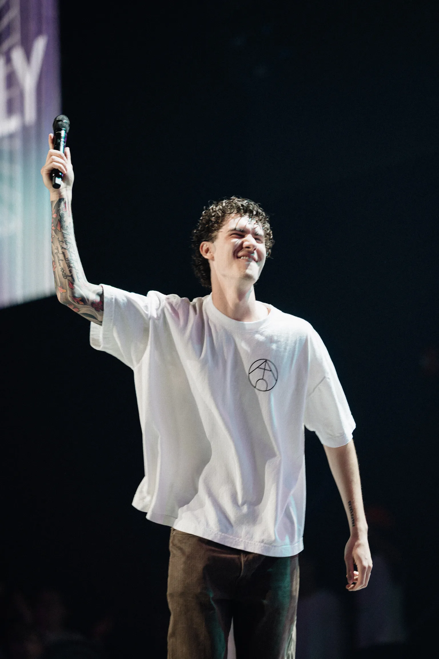 A host raises a microphone overhead while smiling on a dark stage
