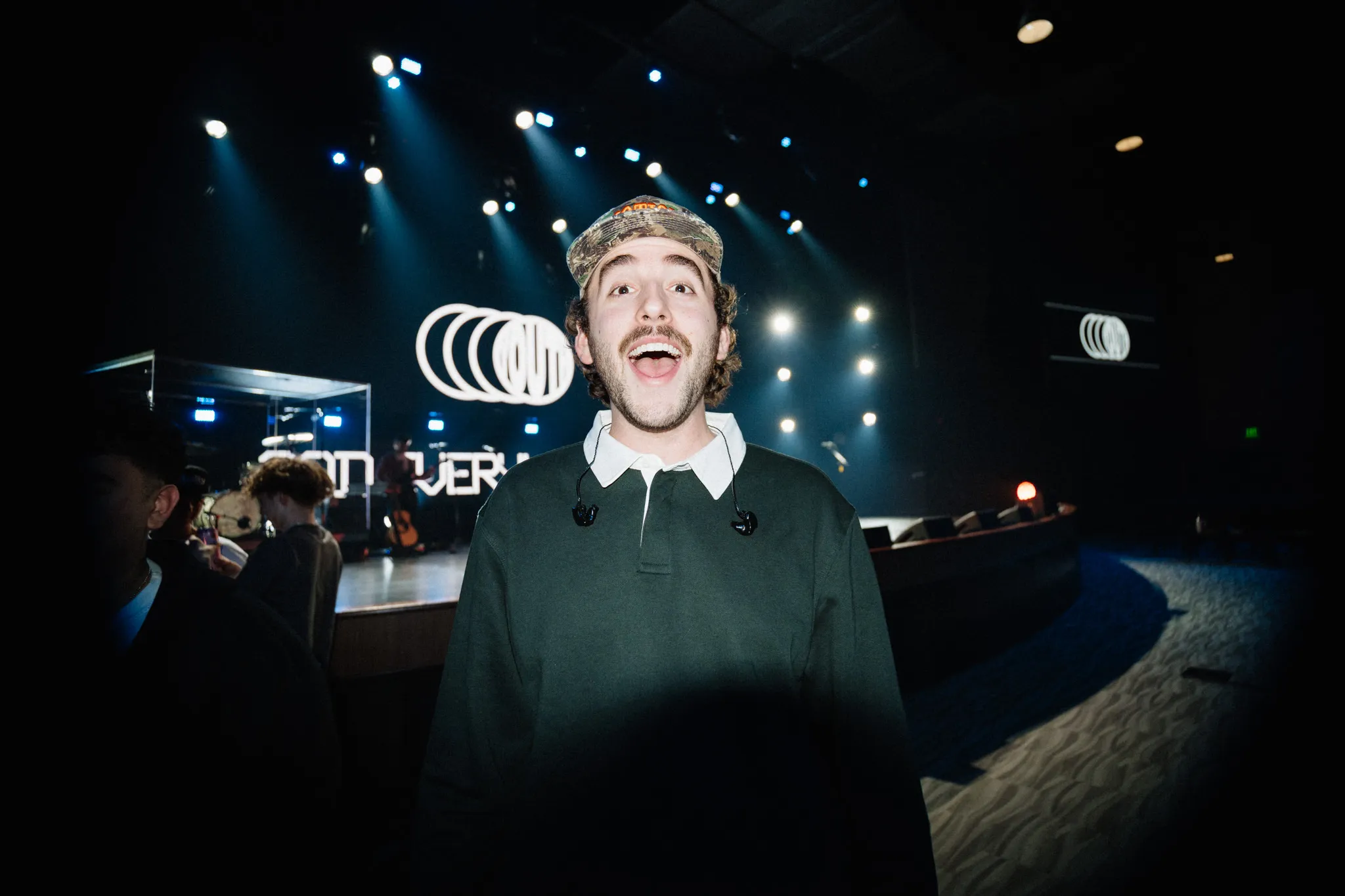 A host in a beanie laughs on stage in front of a lit-up event sign
