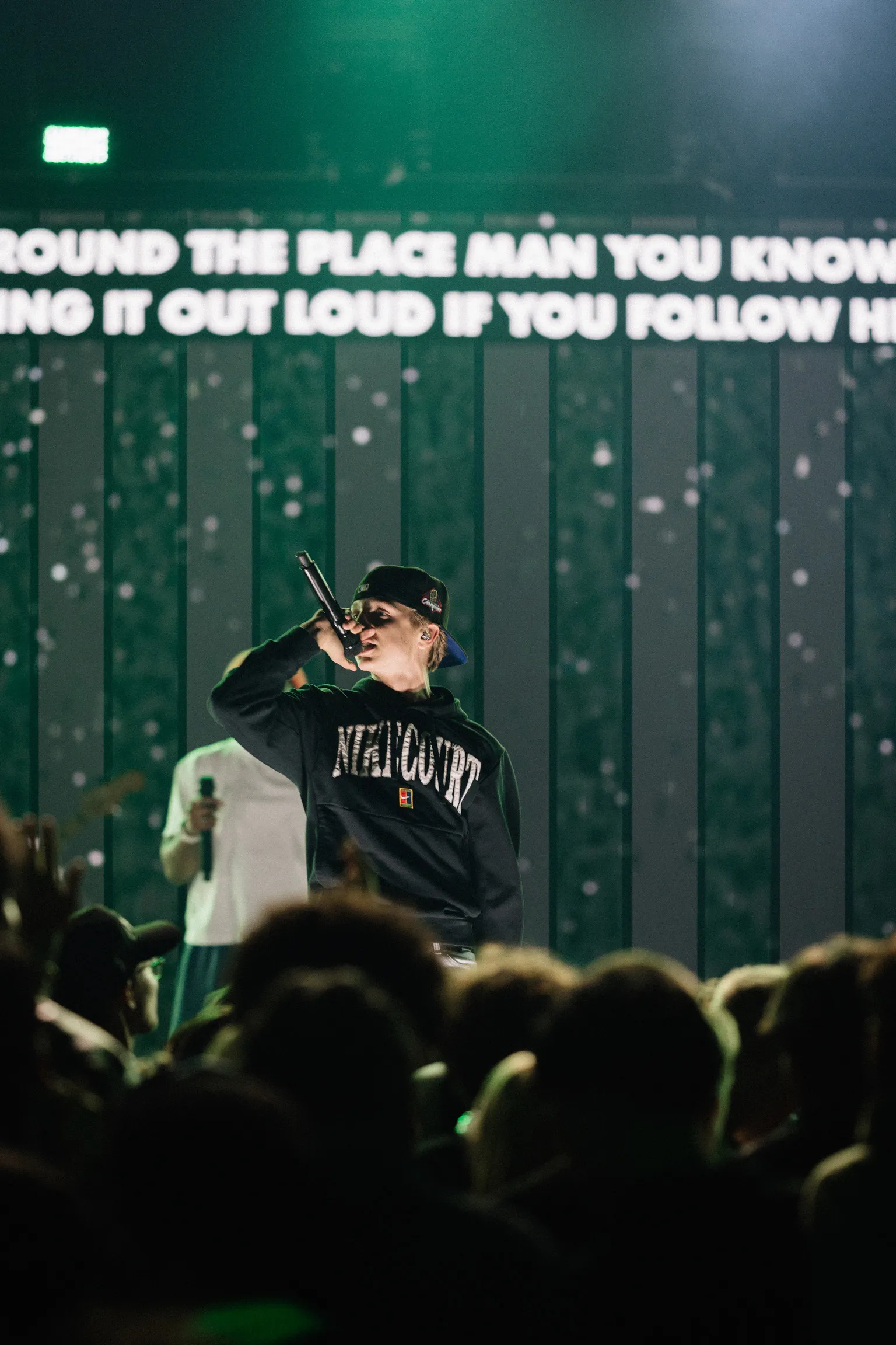 A performer sings into a microphone on stage in front of a green backdrop with lyrics