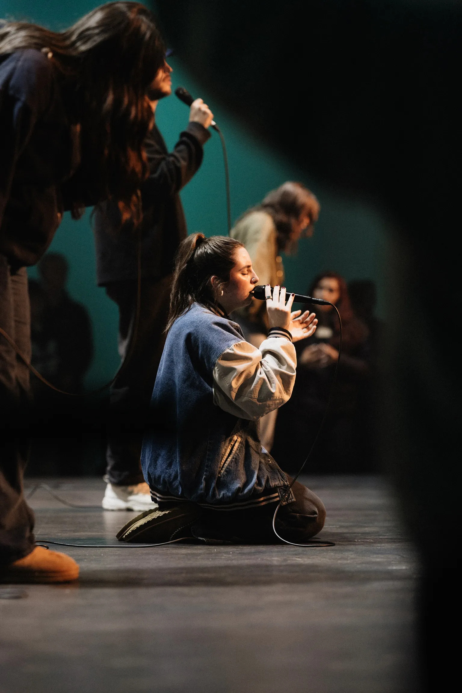 A worship leader kneels on stage singing into a microphone as another vocalist stands behind