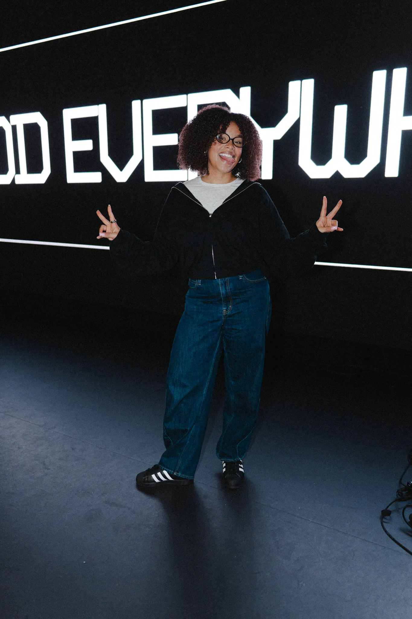 A woman throws up peace signs in front of a lit sign reading "God everywhere"