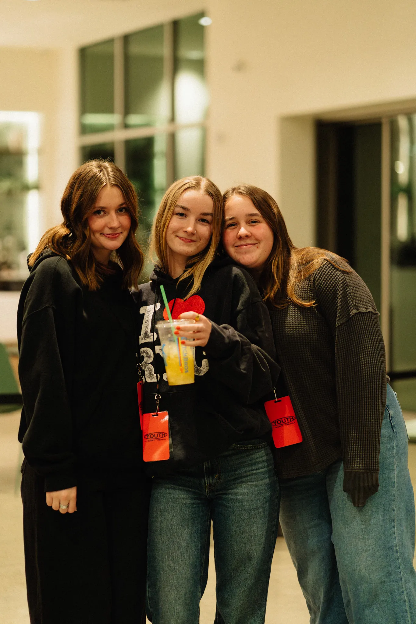 Three friends smile together indoors, one holding a drink with lanyards visible